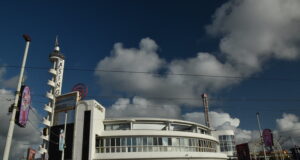 Blackpool Pleasure Beach to Unveil New Aviktas Merch at Free Fashion Show Seaside amusement arcade with a rounded white building, tall sign towers, and colorful lights by the promenade on a sunny day.