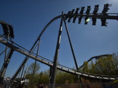 We Love The Swarm at Thorpe Park — And Here’s Why A steel roller coaster loop under a clear blue sky, with riders on a cars train zooming along the track.