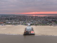 Opinion: Why Britain’s Seaside Piers Still Matter More Than Ever Aerial view of a seaside town at sunset with a pink sky, a long pier extending into calm water, and sandy beach in the foreground.
