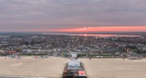 Opinion: Why Britain’s Seaside Piers Still Matter More Than Ever Aerial view of a seaside town at sunset with a pink sky, a long pier extending into calm water, and sandy beach in the foreground.