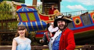 Princesses, Pirates & Pure Family Chaos: Gulliver’s Bank Holiday Takeover Princess in a light blue gown posing with a man in a red pirate coat beside a colorful ship at a theme park.