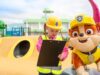 Passholders Get Exclusive First Look at PAW Patrol Land at Chessington Young girl in a yellow hard hat and safety vest checks a clipboard at a colorful playground, beside a large cheerful dog mascot wearing a helmet; bright, playful scene.