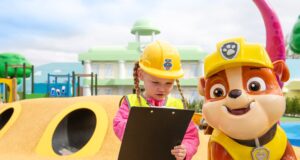 Passholders Get Exclusive First Look at PAW Patrol Land at Chessington Young girl in a yellow hard hat and safety vest checks a clipboard at a colorful playground, beside a large cheerful dog mascot wearing a helmet; bright, playful scene.