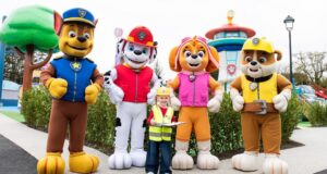 Chessington PAW Patrol Land Tickets: Prices, Dates & How to Save Money Young child in a yellow safety vest and helmet holding a clipboard, posing with five large costumed rescue-dog characters at a park attraction.
