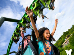 Walibi Holland is fully focusing on the Summer Season and Introduces Summer Saturdays Riders with arms raised on a bright green roller coaster car against a blue sky, showing excitement and motion.