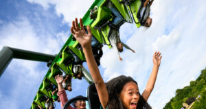 Walibi Holland is fully focusing on the Summer Season and Introduces Summer Saturdays Riders with arms raised on a bright green roller coaster car against a blue sky, showing excitement and motion.