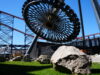 Pleasure Beach Resort release images showing new rides development ahead of opening Close-up of a circular roller coaster wheel and red track structure against a bright blue sky, with large rocks and grass in the foreground