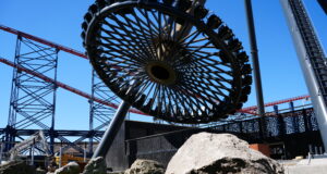 AVIKTAS Preview Day at Blackpool Pleasure Beach: Good Luck Avoiding Spoilers Today Close-up of a circular roller coaster wheel and red track structure against a bright blue sky, with large rocks and grass in the foreground