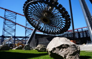 AVIKTAS Preview Day at Blackpool Pleasure Beach: Good Luck Avoiding Spoilers Today Close-up of a circular roller coaster wheel and red track structure against a bright blue sky, with large rocks and grass in the foreground
