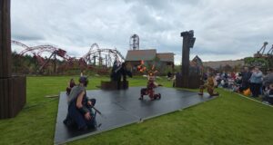 We Attended Valgard’s Opening Day — Here’s Why It Works So Well Fire performers on an outdoor stage display flames while wearing Viking-inspired costumes, with a red roller-coaster track and cheering audience nearby.