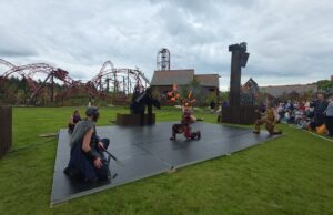 We Attended Valgard’s Opening Day — Here’s Why It Works So Well Fire performers on an outdoor stage display flames while wearing Viking-inspired costumes, with a red roller-coaster track and cheering audience nearby.