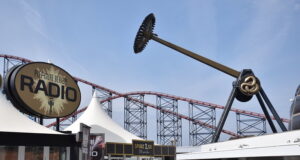 AVIKTAS Is Finally Open at Blackpool Pleasure Beach — And It Was Worth the Wait Large outdoor amusement area with a circular 'Pleasure Beach Radio' sign and red roller coaster tracks in the background.