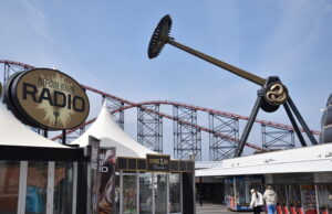 AVIKTAS Is Finally Open at Blackpool Pleasure Beach — And It Was Worth the Wait Large outdoor amusement area with a circular 'Pleasure Beach Radio' sign and red roller coaster tracks in the background.