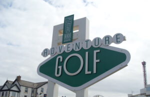 Save 50% on Adventure Golf at Blackpool Pleasure Beach Large green-and-white sign reading 'Adventure Golf' at a seaside resort, with a 'Pleasure Beach' emblem above.