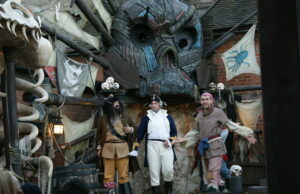 We Can’t Be the Only Ones Still Missing the Pirate Show in Mutiny Bay Three performers in pirate costumes center stage in a steampunk pirate set with a large mechanical creature face above and skulls nearby.