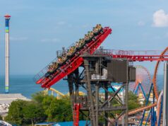 Cedar Point’s New “Morning Rush” Event: Is It Worth the Early Wake-Up? Red roller coaster climbs upward with riders wearing neon vests against a seaside park backdrop and blue sky.