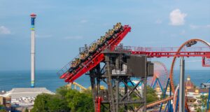 Cedar Point’s New “Morning Rush” Event: Is It Worth the Early Wake-Up? Red roller coaster climbs upward with riders wearing neon vests against a seaside park backdrop and blue sky.