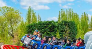 PAW Patrol Opens at Chessington World of Adventures: Everything You Need to Know About the UK’s First Pup-Themed Land Riders on a blue roller-coaster train outdoors, smiling, with a mascot in the front car and trees in the background.
