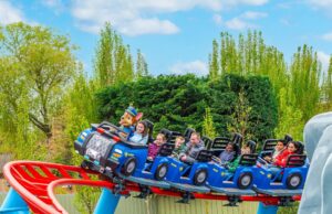 PAW Patrol Opens at Chessington World of Adventures: Everything You Need to Know About the UK’s First Pup-Themed Land Riders on a blue roller-coaster train outdoors, smiling, with a mascot in the front car and trees in the background.