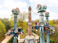 Drop tower attraction Hooghmoed opens at Efteling Riders on a tall, steampunk-inspired roller coaster with turquoise towers, arms raised, soaring above trees on a sunny day.
