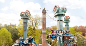 Drop tower attraction Hooghmoed opens at Efteling Riders on a tall, steampunk-inspired roller coaster with turquoise towers, arms raised, soaring above trees on a sunny day.