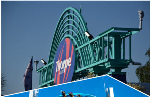 Could Cedar Point’s “Morning Rush” Concept Work at UK Theme Parks? Entrance sign at Cedar Point with teal arches and a roller coaster structure against a blue sky as riders zoom by above the track.