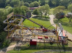 What’s Happening at UK Theme Parks This Week (Week of 25 May 2026) Elevated view of a bright yellow roller coaster with purple rails and a 'Barnyard Whirl' sign, set among green trees and park paths.