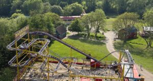 What’s Happening at UK Theme Parks This Week (Week of 25 May 2026) Elevated view of a bright yellow roller coaster with purple rails and a 'Barnyard Whirl' sign, set among green trees and park paths.