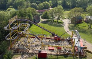 Pleasurewood Hills Owner Confirms Wipeout Plans and Four New Rides for 2027 Elevated view of a bright yellow roller coaster with purple rails and a 'Barnyard Whirl' sign, set among green trees and park paths.