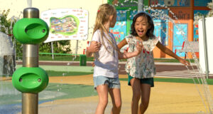 A Refreshing Summer at Futuroscope Xperiences Two girls laugh and play in a colorful water-spray playground, holding hands as water jets spray around them.