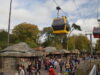 Alton Towers Skyride Reopens This Saturday Yellow gondola cabin network glides above a crowded outdoor market area in a park, with rustic huts and trees nearby.