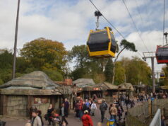 Alton Towers Skyride Reopens This Saturday Yellow gondola cabin network glides above a crowded outdoor market area in a park, with rustic huts and trees nearby.