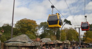 Alton Towers Skyride Reopens This Saturday Yellow gondola cabin network glides above a crowded outdoor market area in a park, with rustic huts and trees nearby.