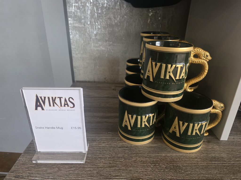 Green ceramic AVIKTAS mug with a gold snake-shaped handle, stacked on a wooden shelf beside a display card.