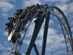 Which UK Theme Park Has the Strongest Coaster Line-Up? Roller coaster train arcs over a large steel loop with riders secured in their seats against a blue sky backdrop.