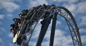 Which UK Theme Park Has the Strongest Coaster Line-Up? Roller coaster train arcs over a large steel loop with riders secured in their seats against a blue sky backdrop.