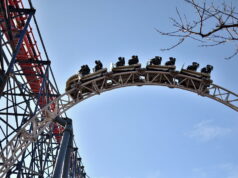 What’s Happening at UK Theme Parks This Week (Week of 22 June 2026) Roller coaster train racing along a high curved track against a clear blue sky, with a tree branch in the corner.