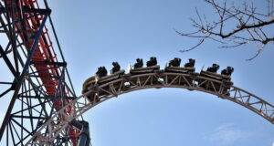 What’s Happening at UK Theme Parks This Week (Week of 22 June 2026) Roller coaster train racing along a high curved track against a clear blue sky, with a tree branch in the corner.