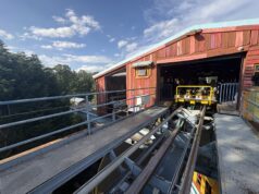 Behind the Scenes on The Wave at Drayton Manor – Inside the Station & Lift Hill Outdoor amusement ride station with colorful wooden facade and yellow ride cars on exposed tracks against a blue sky and trees in the background.