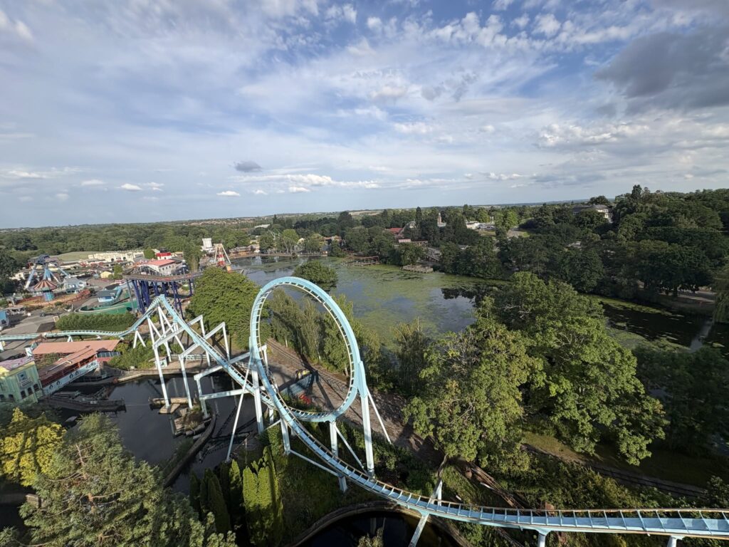 Aerial view of a blue roller coaster looping through a park with trees, a river, and buildings in the background.