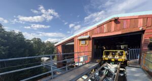 Behind the Scenes on The Wave at Drayton Manor – Inside the Station & Lift Hill Outdoor amusement ride station with colorful wooden facade and yellow ride cars on exposed tracks against a blue sky and trees in the background.