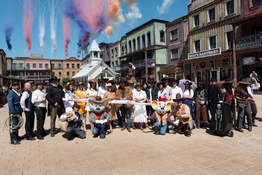 Group of people in cowboy and period costumes posing for a photo on a Western town street with colorful smoke plumes in the sky behind them using a sunny day setting.
