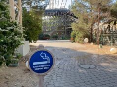 Europa-Park Announces New Smoking Restrictions Starting June 2026 Blue round sign reading 'Raucherbereich Smoking Area' with cigarette graphic in foreground; outdoor park path and roller coaster in background.