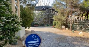 Europa-Park Announces New Smoking Restrictions Starting June 2026 Blue round sign reading 'Raucherbereich Smoking Area' with cigarette graphic in foreground; outdoor park path and roller coaster in background.