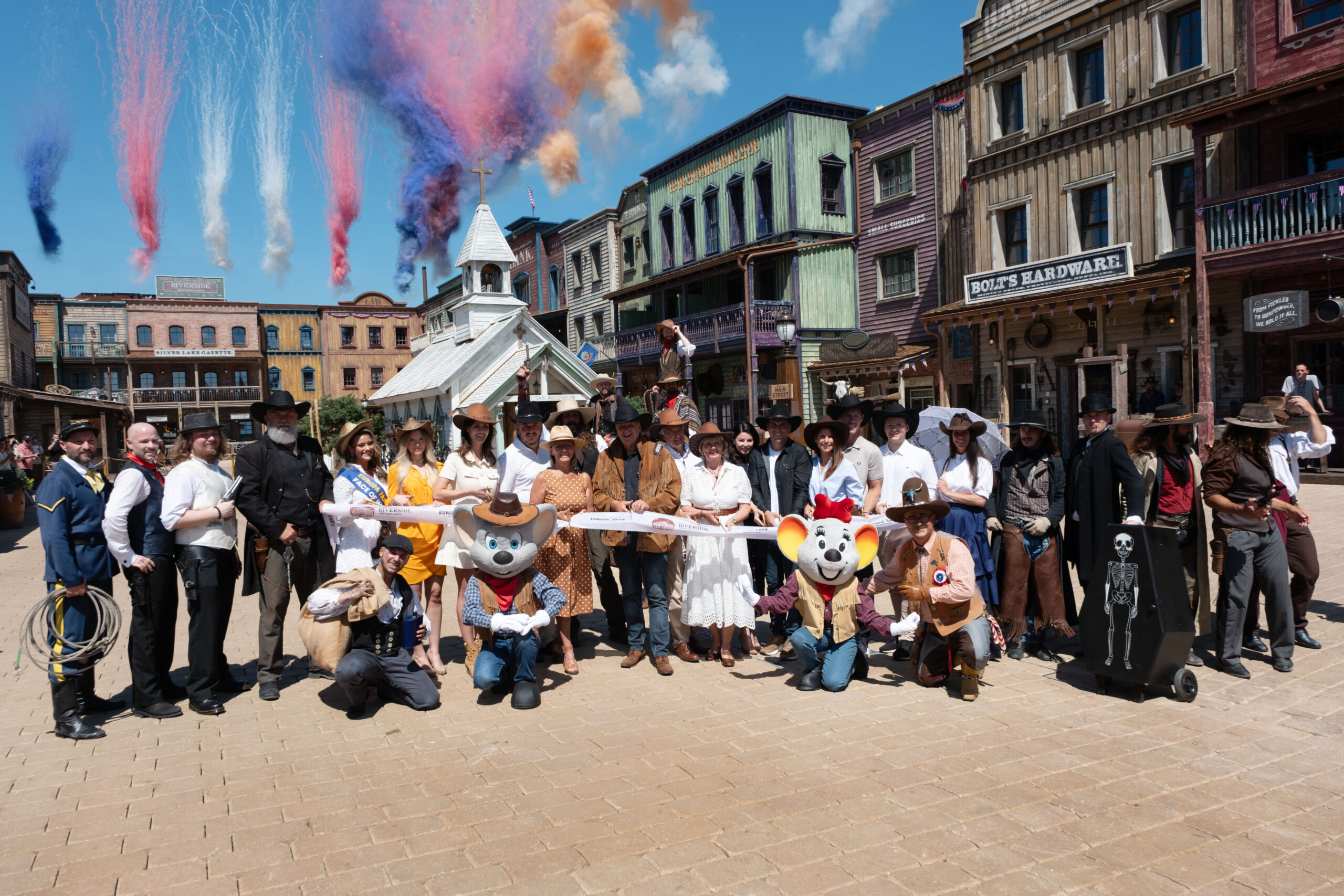 Riverside Western Lodge Opens at Europa-Park Resort’s Silver Lake City Group of people in cowboy and period costumes posing for a photo on a Western town street with colorful smoke plumes in the sky behind them using a sunny day setting.
