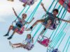 Great British Summer Savings at Harbour Park Children joyfully ride on a colorful carnival swing ride high in the air against a bright blue sky.