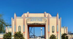 Celebrate 30 Years: Movie Park Germany’s Summer Lineup Entrance to Movie Park Germany with pastel Art Deco arches and turquoise accents, flanked by palm trees against a blue sky.",