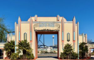 Celebrate 30 Years: Movie Park Germany’s Summer Lineup Entrance to Movie Park Germany with pastel Art Deco arches and turquoise accents, flanked by palm trees against a blue sky.",