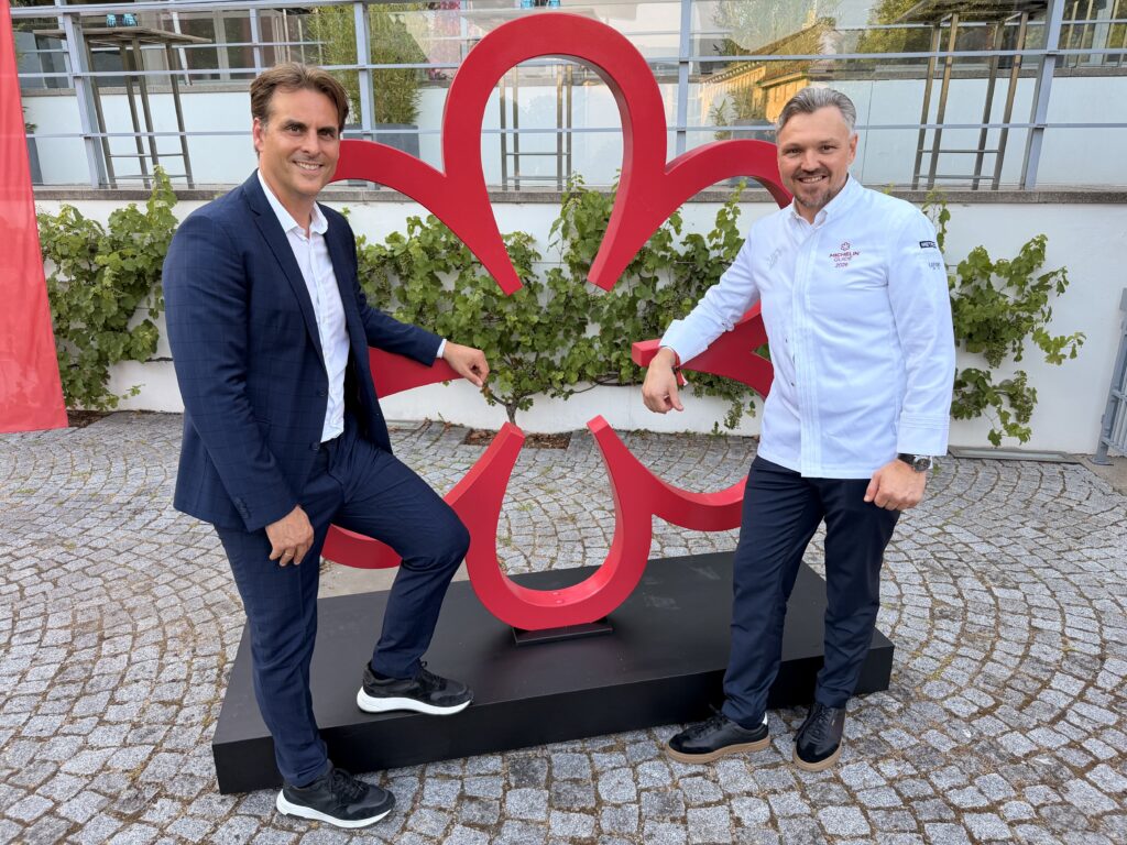 Two men in smart outfits pose next to a large red abstract sculpture outdoors, standing on a black pedestal.