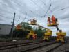 Network Rail to Deliver New Wixams Station Supporting Universal United Kingdom Resort Railway workers in orange high-visibility suits on yellow elevated platforms inspect overhead power lines above tracks under a cloudy sky.